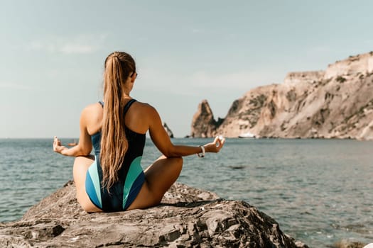 Yoga on the beach. A happy woman meditating in a yoga pose on the beach, surrounded by the ocean and rock mountains, promoting a healthy lifestyle outdoors in nature, and inspiring fitness concept