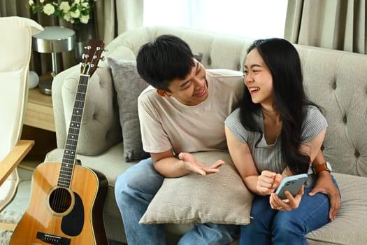 Cheerful young Asian couple using smartphone on comfortable couch in living room.
