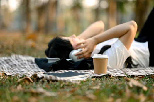 A paper cup of coffee on checkered blanket near relaxed young woman listening to music with headphone.