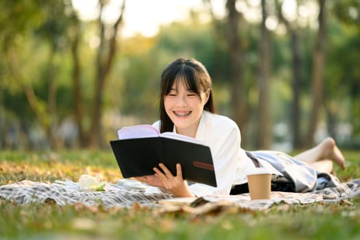 Cheerful young Asian woman lying down on green grass and reading a book.