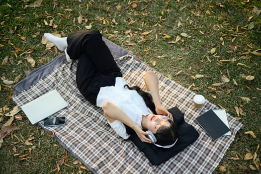Overhead view of young woman lying on lawn resting with closed eyes and listening music in headphone.