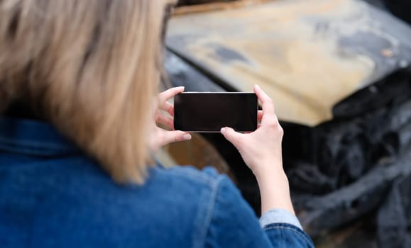 Insurance agent woman photographs burned-out car on smartphone. Transport insurance and insurance payment after fire