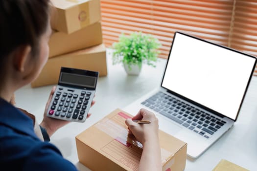 Entrepreneur using calculator in her hand, calculating financial expense at home office, online market packing box delivery.