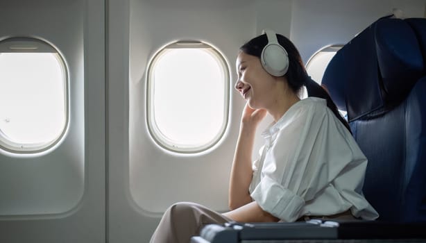 Young woman wearing headphones listening to music during travel, sitting near window in first class on airplane during flight, travel and business concept.