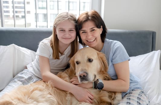 Cheerful Mom And Daughter Sit In Bed With Golden Retriever Dog, Enjoying Morning At Home