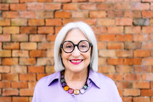 portrait of a beautiful senior woman with glasses smiling happy looking at camera with a brick wall in the background, concept of happiness of elderly people and active lifestyle