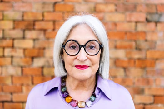 portrait of a beautiful senior woman with glasses looking at camera with a brick wall in the background, concept of happiness of elderly people and active lifestyle