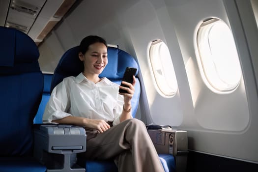 Young Asian woman checks business news on mobile phone, sitting near window in first class on airplane during flight, travel and business concept.