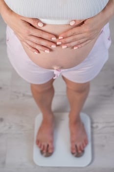 Top view of a pregnant woman in home clothes standing on an electronic scale and holding her hands on her tummy