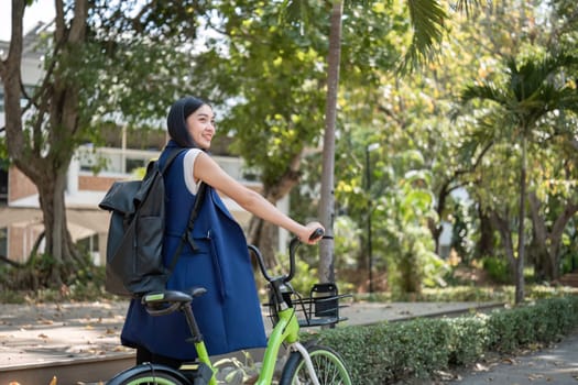 A young Asian woman commutes to work by cycling in a green city, carrying a backpack and using a reusable drinking cup to avoid harming the environment..