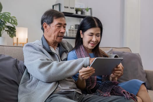 A senior couple in their 60s spends their free time sitting on a tablet, relaxing and watching entertainment programs, relaxing happily together on the sofa in the living room..