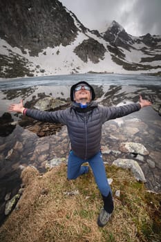 Happy active cheerful tourist in mountain sunglasses and a down jacket, joyful smiles expressively rejoices against the backdrop of a mountain lake high in the mountains.