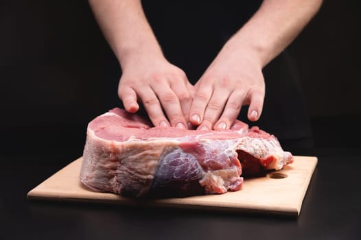 A man massages a fresh piece of beef with his hands on a wooden cutting board. Close up on black background.
