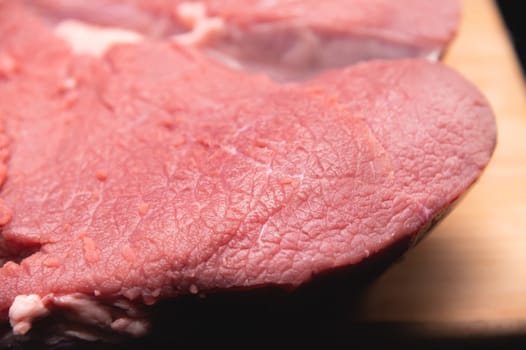 A piece of raw meat lies on a cutting board on a dark background. Close-up of beef meat fibers.