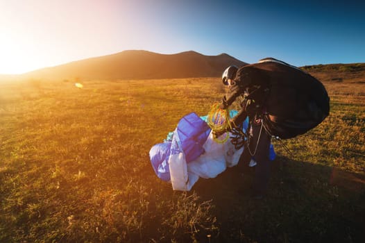 A young male paraglider at sunset in the mountains collects his paraglider to take it to the starting point. Paragliding sport.