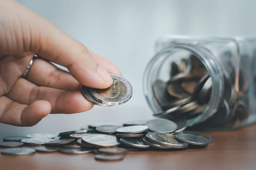 Hand dropping coins into a glass jar overflowing with coins symbolizes the concept of saving money and financial planning.