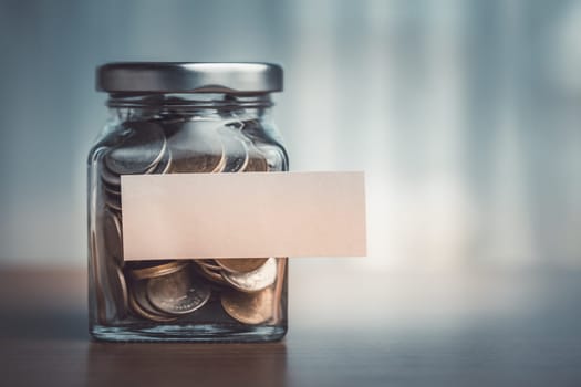 Coins stored in a glass jar with a blank paper label placed on a wooden table, suggesting a simple yet practical method of saving and organizing spare change or funds for future use.