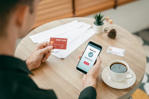 Young man sitting at table using online payment app and digital wallet on smartphone to pay with credit card. E commerce shopping and modern purchasing via mobile internet. Unveiling