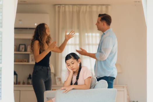 Annoyed and unhappy young girl sitting on sofa trapped in middle of tension by her parent argument in living room. Unhealthy domestic lifestyle and traumatic childhood develop to depression.Synchronos