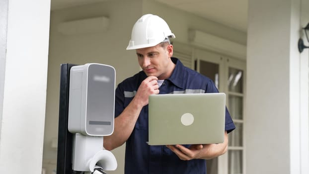 Qualified technician working on home EV charging station installation, making troubleshooting and configuration setup on charging system with laptop for EV at home. Synchronos