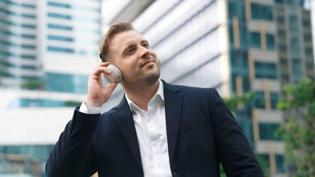 Close up of male leader listen headphone and dancing to relaxing music with cheerful and motivation. Skilled project manager enjoy to listen relaxed song while hold smart phone at urban city. Urbane.