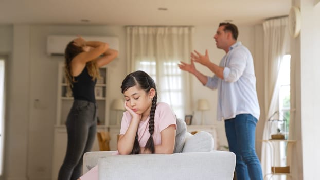 Annoyed and unhappy young girl sitting on sofa trapped in middle of tension by her parent argument in living room. Unhealthy domestic lifestyle and traumatic childhood develop to depression Synchronos