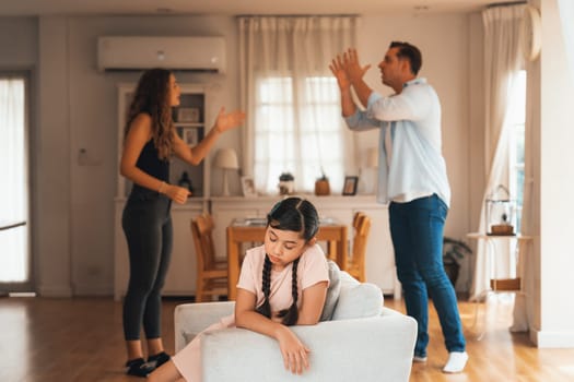 Annoyed and unhappy young girl sitting on sofa trapped in middle of tension by her parent argument in living room. Unhealthy domestic lifestyle and traumatic childhood develop to depression.Synchronos