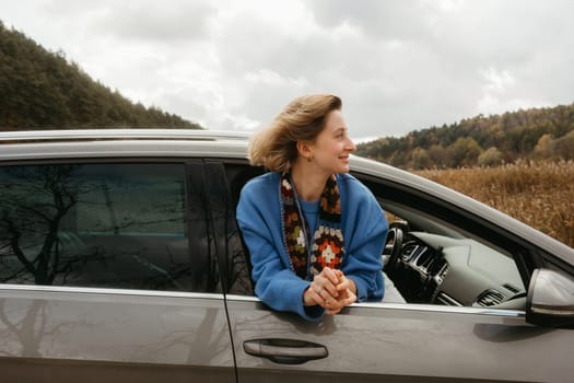 A woman leans out of car window during road trip in autumn, the wind blows her hair