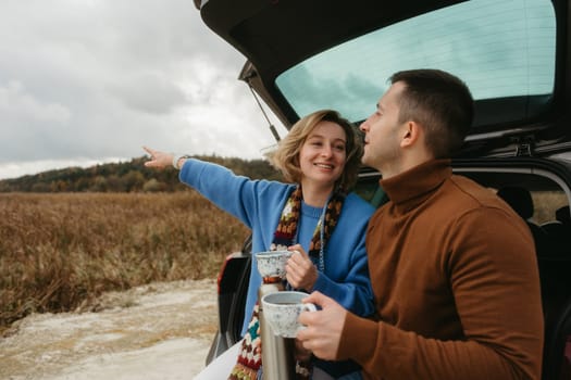 Happy young woman and man sitting in the open trunk of car while traveling in autumn, road trip concept