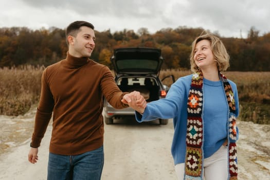 Smiling couple holding hands, strolling down country road with car on the background, happy man and woman on roadtrip