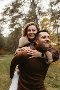 Adult couple having fun in the autumn park outdoors, happy man is carrying woman on his back in the woods
