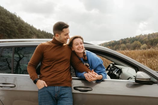 Happy man leaning to car and holding hands of cheerful woman that sitting inside, adult couple enjoying road trip in autumn