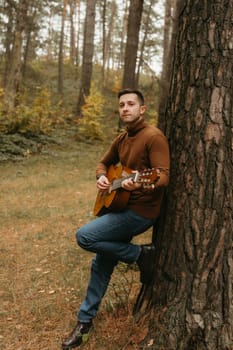 Caucasian adult man plays guitar leaning on tree in the autumn park