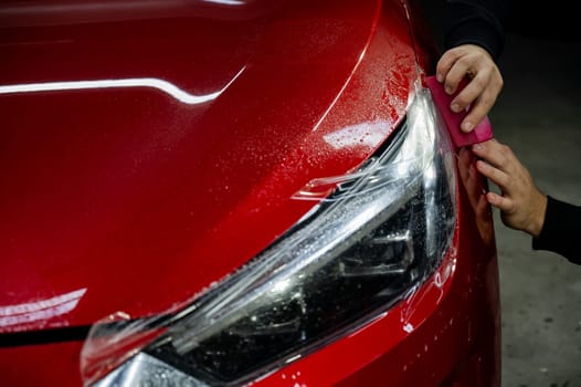 The master applies vinyl film to the headlight of a red car. Closeup view on worker detailer hand smoothing with a scraper protective film