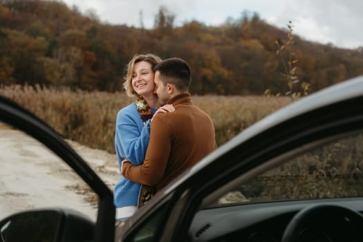 Embracing man and a woman stand next to a car on dirt road and smiling, couple enjoying roadtrip