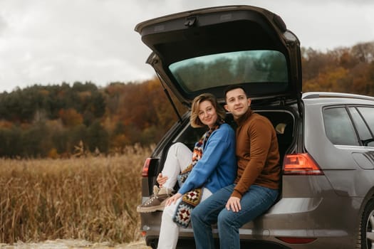Happy young woman and man sitting in the open trunk of car while traveling in autumn and looking away, road trip concept