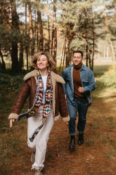 Happy couple is having fun in the middle of the forest, smiling woman is running with her husband in an autumn park