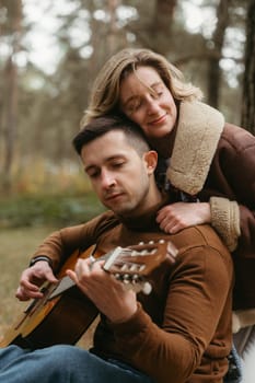 Happy couple spending time together in autumn park, a man playing guitar while woman hugs him