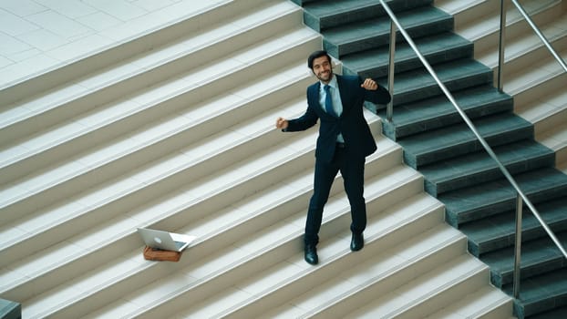 Professional business man standing at stair while moving to music from headphone. Skilled project manager dancing while listening relax sound with laptop placed on stairs. Modern dance. Exultant.