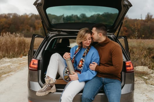 Happy adult couple sitting in the open trunk of car while traveling in autumn, road trip concept, man embracing woman