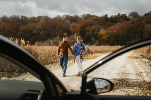 Happy man and a woman are walking down a country road next to their car holding hands, adult couple on road trip in autumn