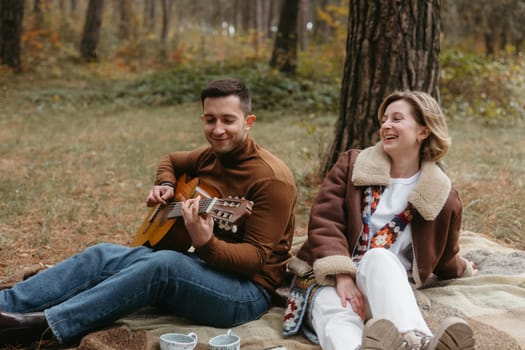 A man is playing a guitar for a happy woman while they are sitting on blanket in the autumn woods