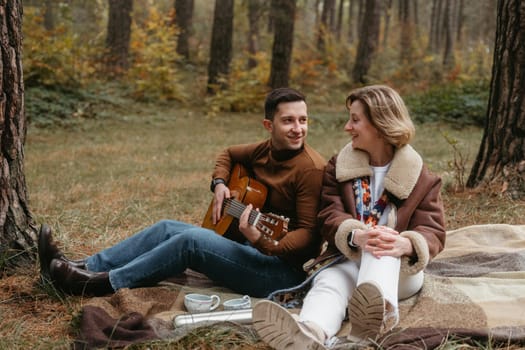 A cheerful man is playing a guitar for a happy woman while they are sitting on blanket in the autumn woods