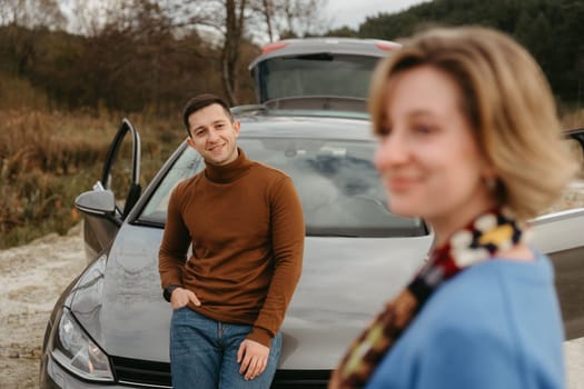 Couple enjoying roadtrip, man leaning on the car hood with woman in front of him on country road and smiling