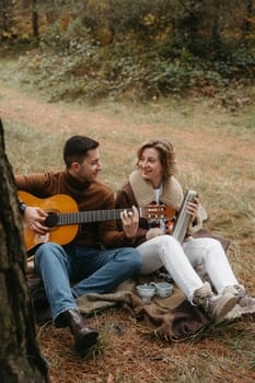 Man is playing guitar for happy woman, adult caucasian couple sitting on blanket in autumn park