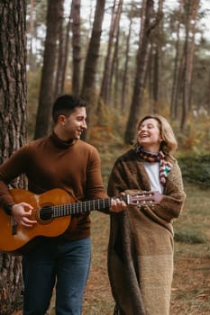 A man in jeans is playing a guitar for a woman covered in plaid. They are surrounded by trees and a natural landscape, both smiling