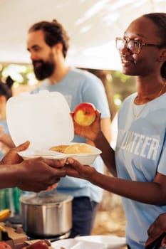 Close-up shot of generous black woman working in community food bank, serving free meals for the needy. Volunteering team of charity workers helping and sharing nutritrious items to homeless people.