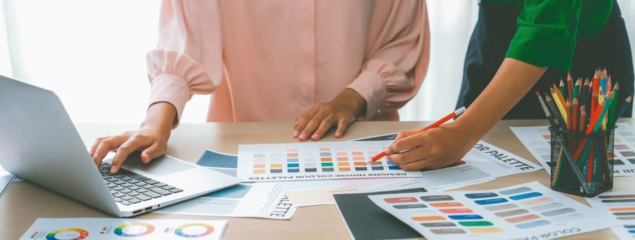 Cropped image of two female young beautiful graphic designer choose the color from color palate by using laptop on table with work tools and equipments scatter around at modern office. Variegated.