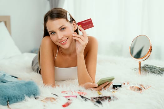 Young woman lying on the bed using online payment app and digital wallet on smartphone to pay with credit card. E commerce shopping and modern purchasing via mobile internet. Unveiling