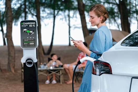 Outdoor adventure and family vacation camping at sea travel by eco friendly car. Woman or mother check car's battery with smartphone while charging EV car frin charging station in campsite. Perpetual